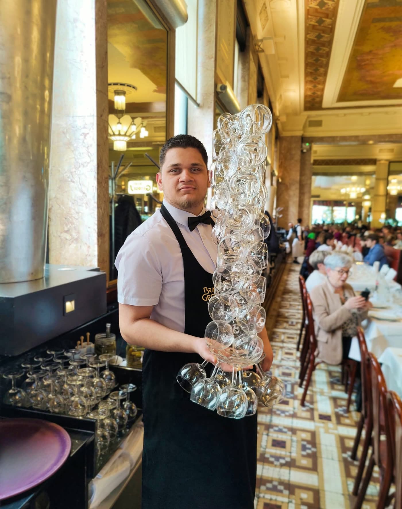 Serveur en tablier tenant une haute pile de verres à vin en équilibre près du bar — Brasserie Georges