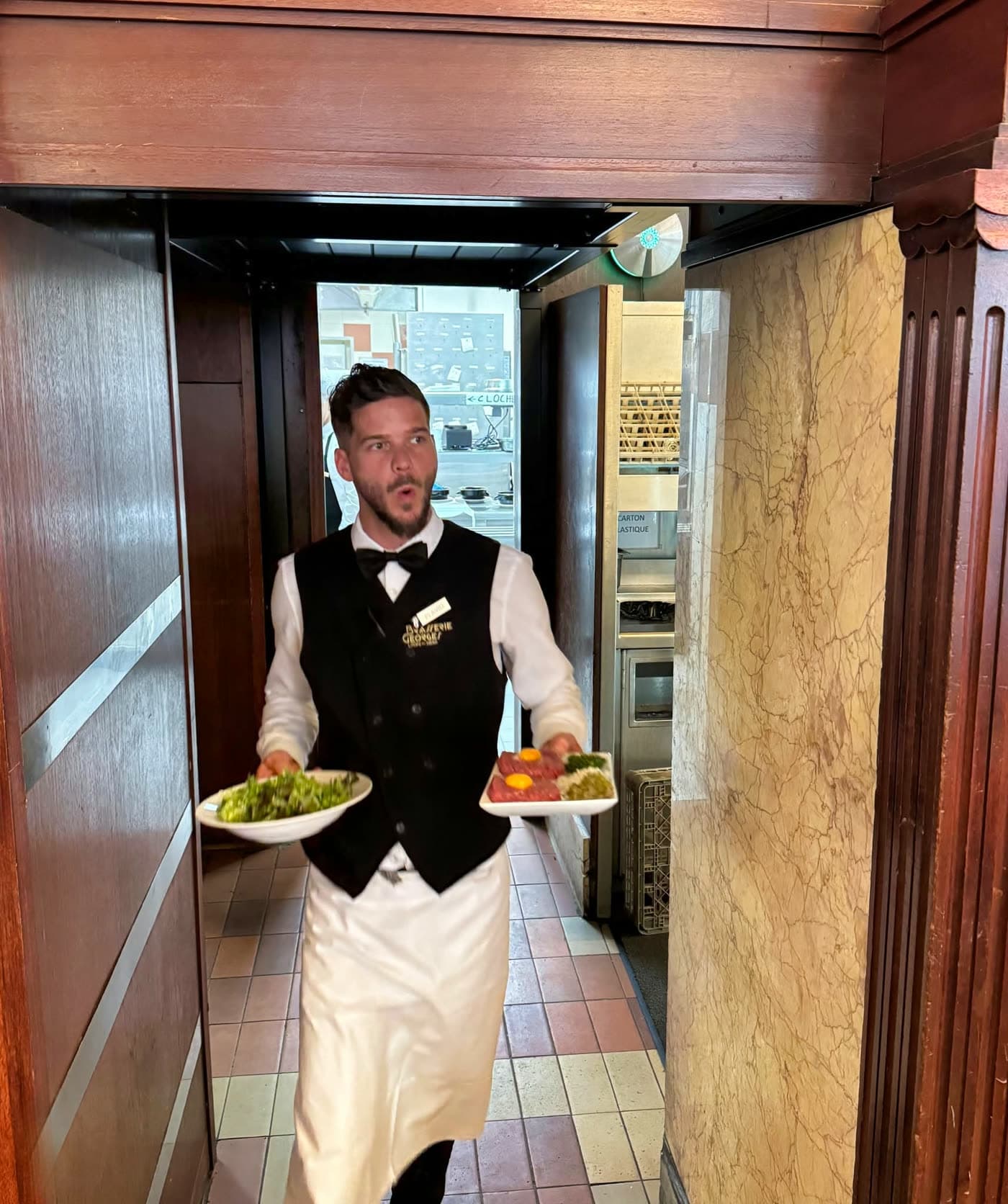 Serveur en gilet et nœud papillon traverse un couloir lambrissé avec deux assiettes — Brasserie Georges