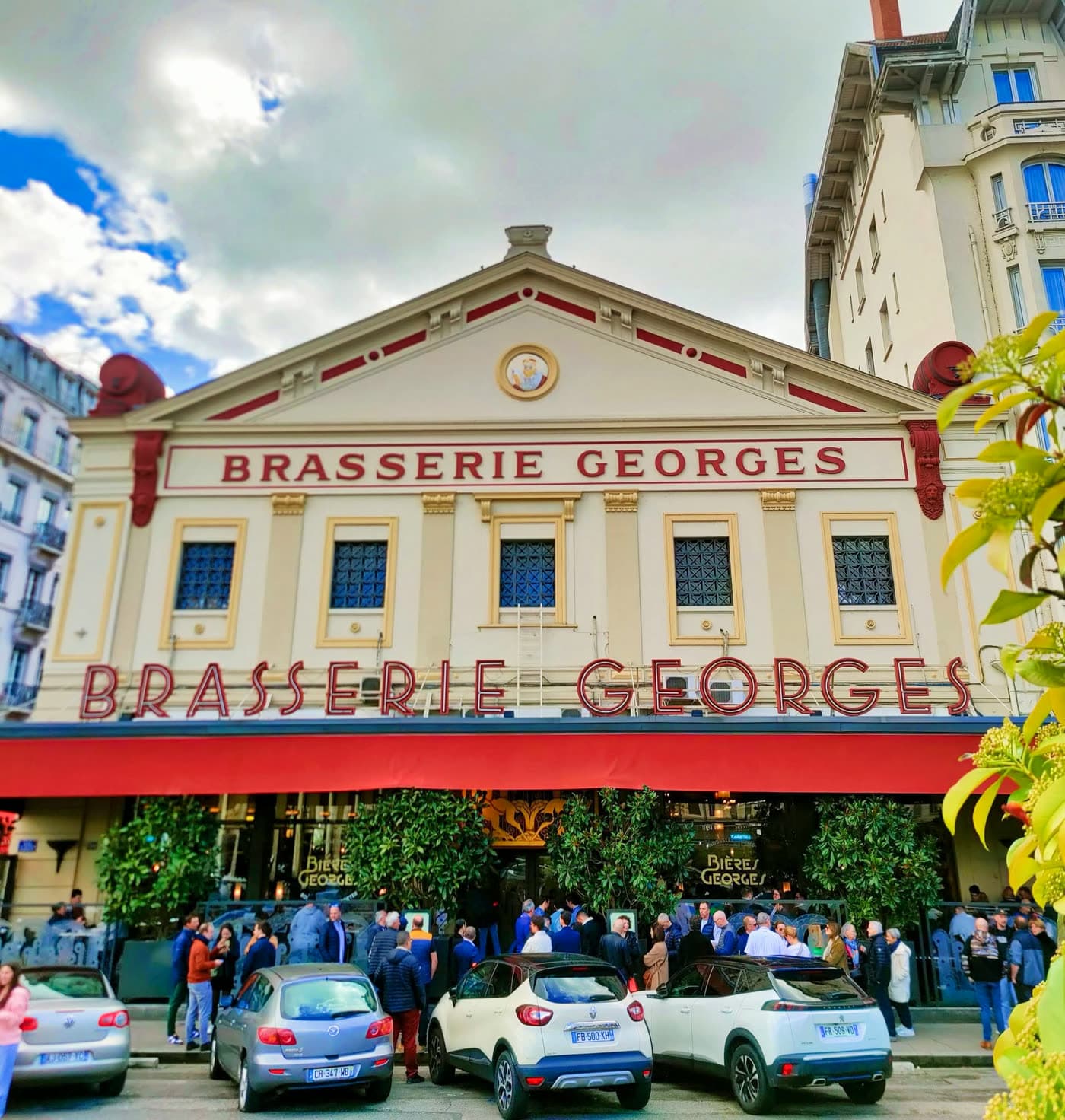 Façade classique Brasserie Georges de jour, enseignes rouges, voitures garées devant