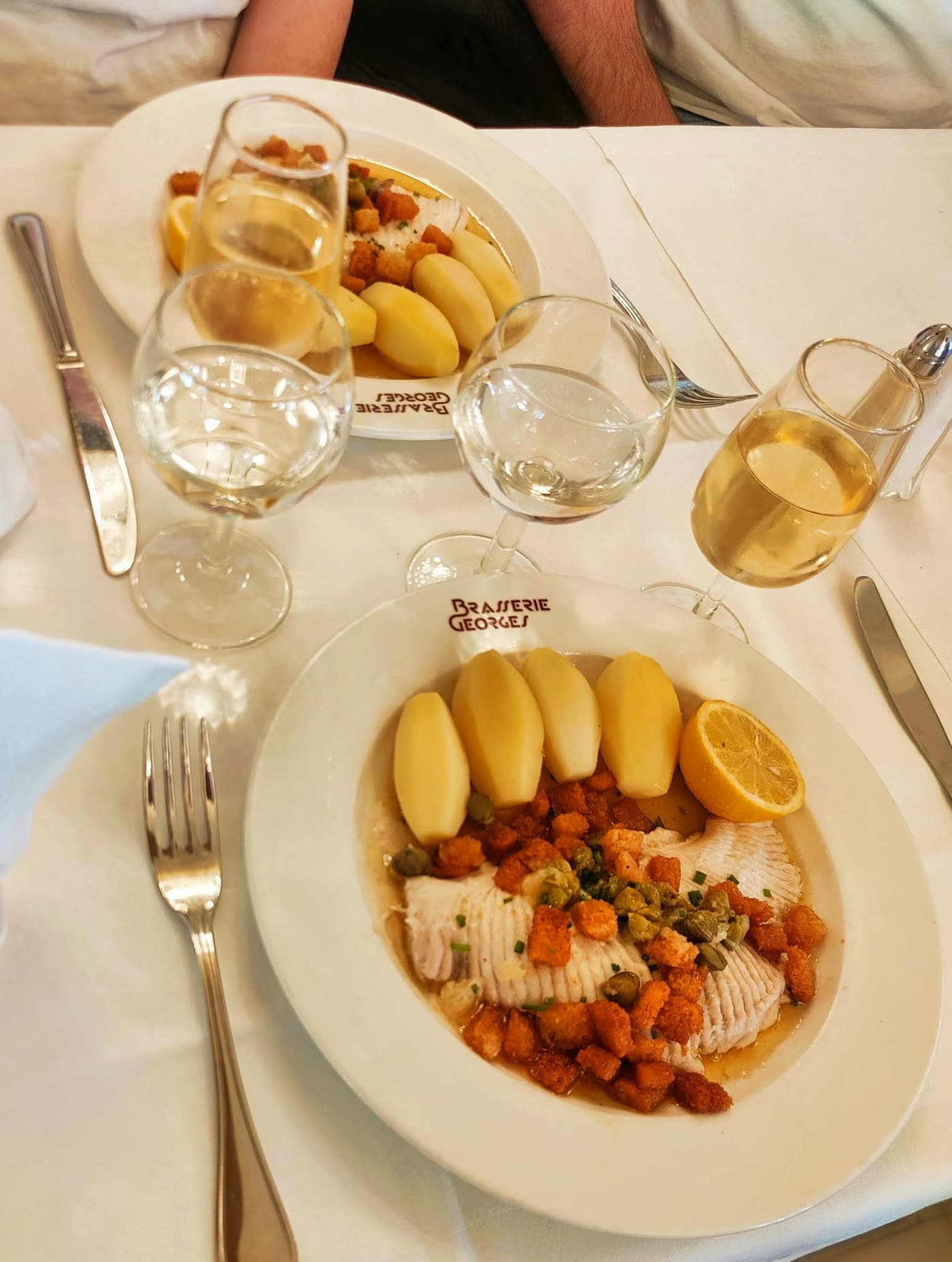 Deux assiettes de filets de poisson pané, pommes vapeur, croûtons et citron, verres de vin — Brasserie Georges