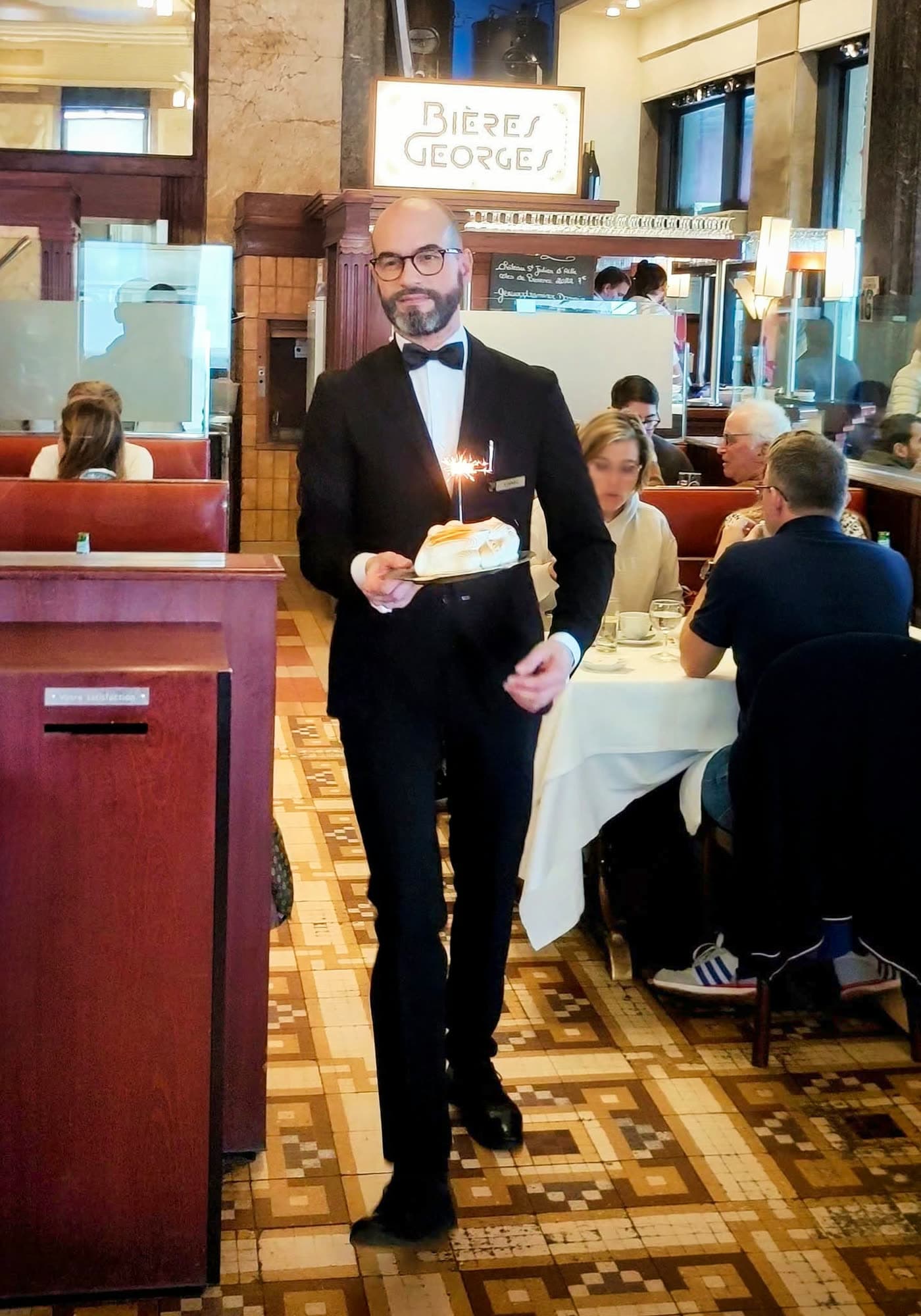 Maître d'hôtel en smoking porte un dessert avec bougie allumée, salle en arrière-plan sous enseigne Bières Georges — Brasserie Georges