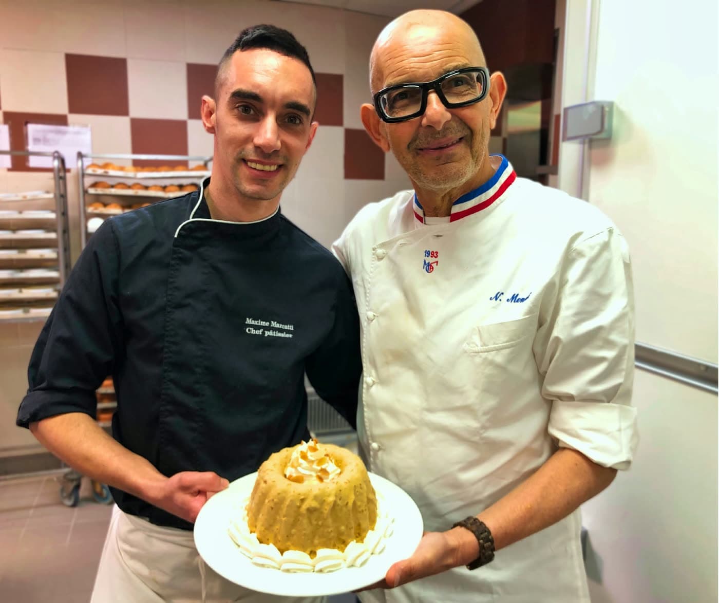 Deux pâtissiers en cuisine posent avec un gâteau glacé au caramel, col tricolore MOF pour l'un — Brasserie Georges