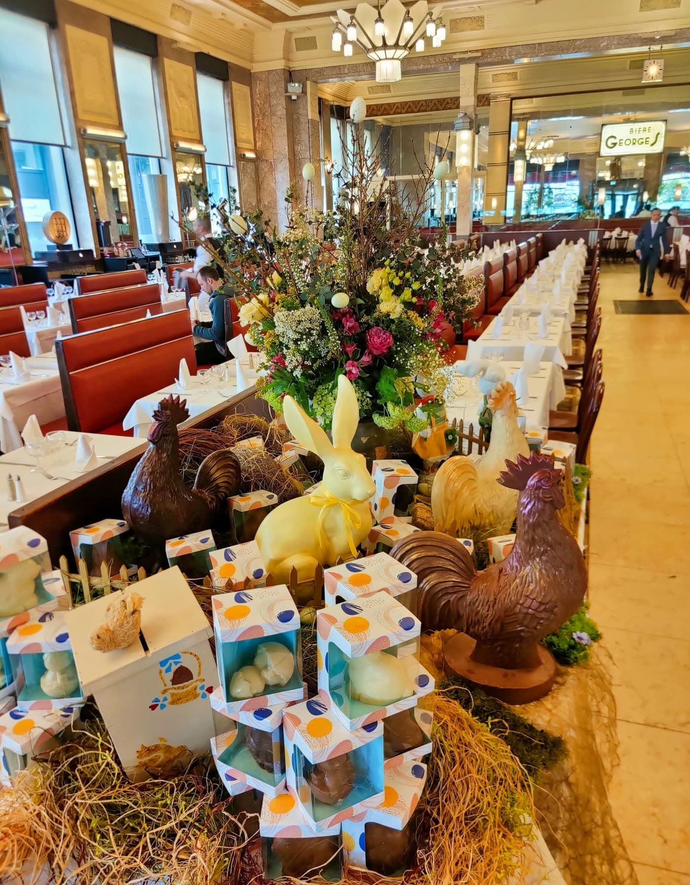 Longue tablée dressée avec décor de Pâques en chocolat et fleurs, enseigne Georges au fond — Brasserie Georges