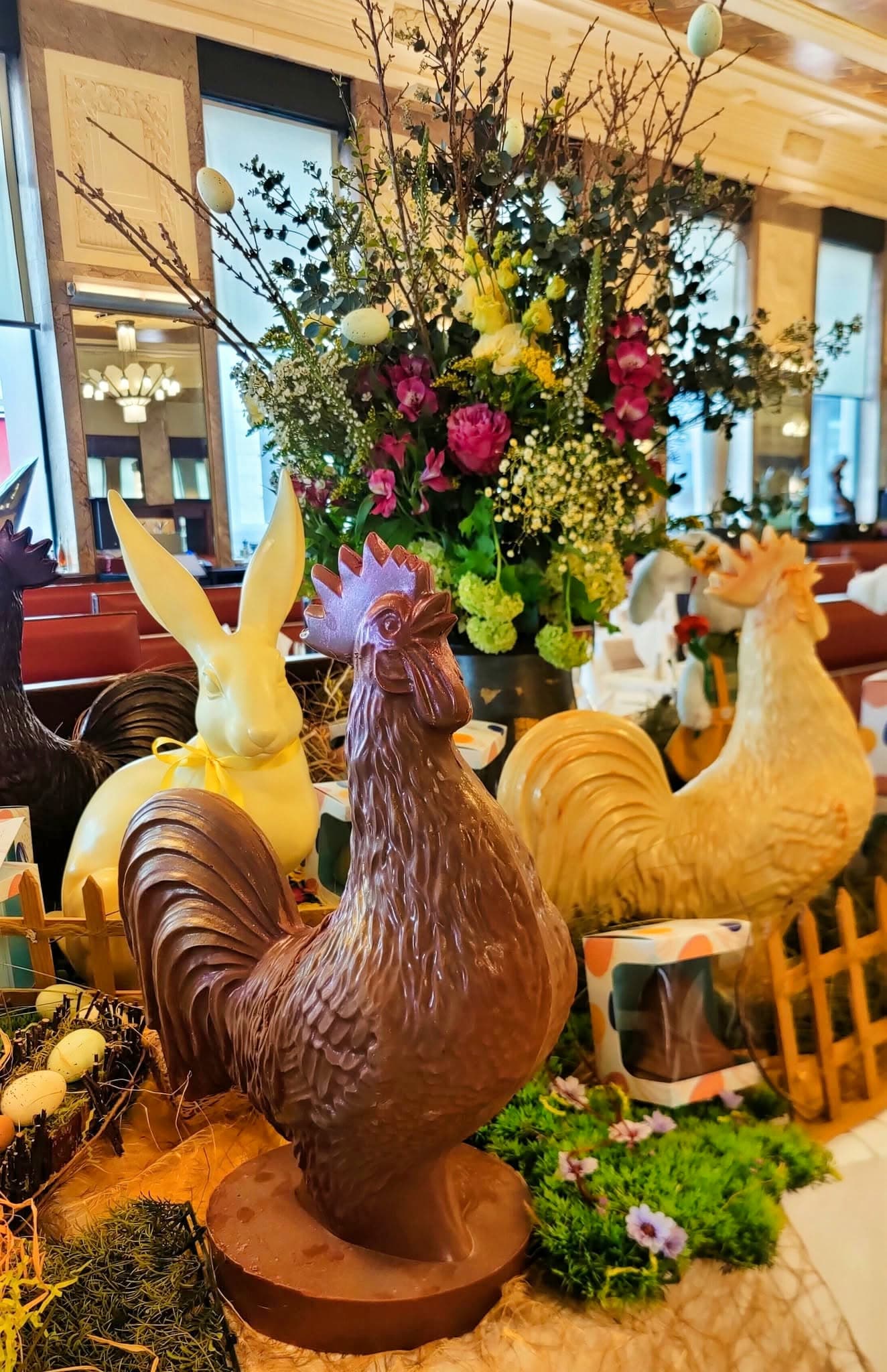 Gros plan sur un coq en chocolat entouré de fleurs, salle floue en arrière-plan — Brasserie Georges