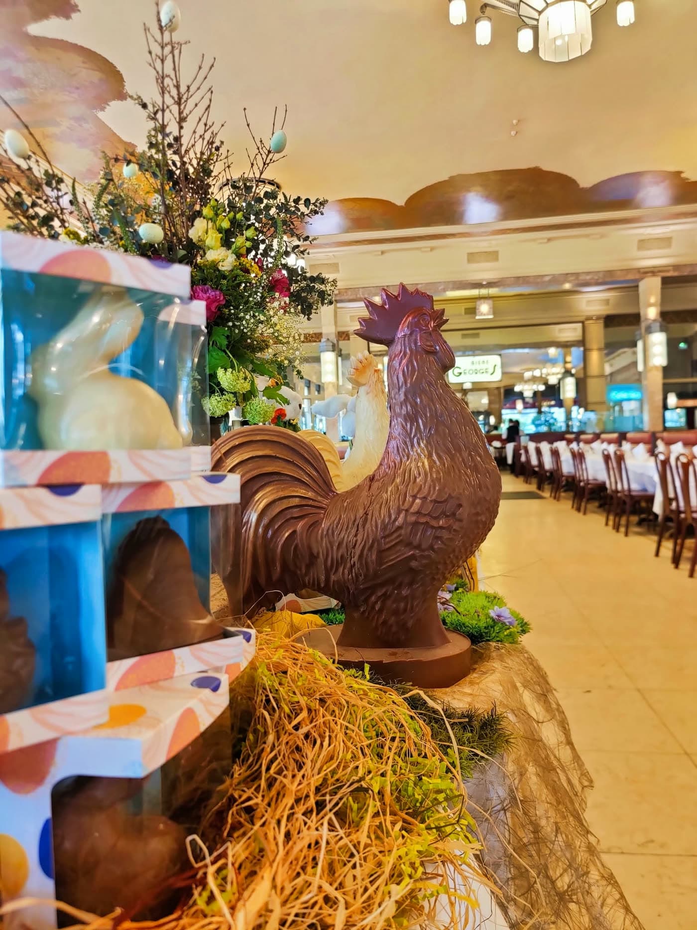 Coq en chocolat et boîtes cadeaux au premier plan, salle avec enseigne Georges floue en fond — Brasserie Georges