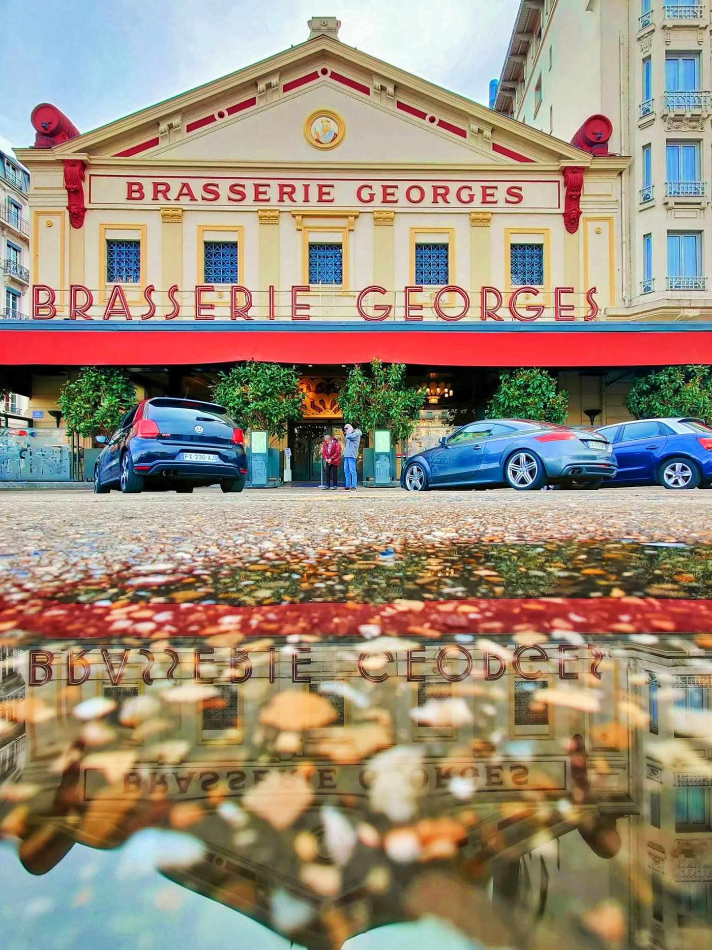 Façade Brasserie Georges avec reflet dans une flaque d'eau au sol, voitures garées devant
