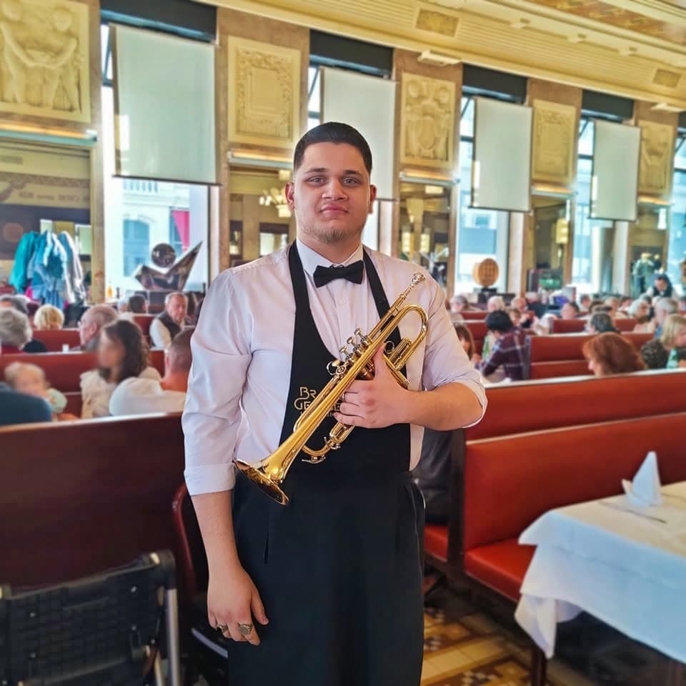 Jeune musicien en gilet noir pose avec sa trompette dans la salle du restaurant remplie de clients — Brasserie Georges