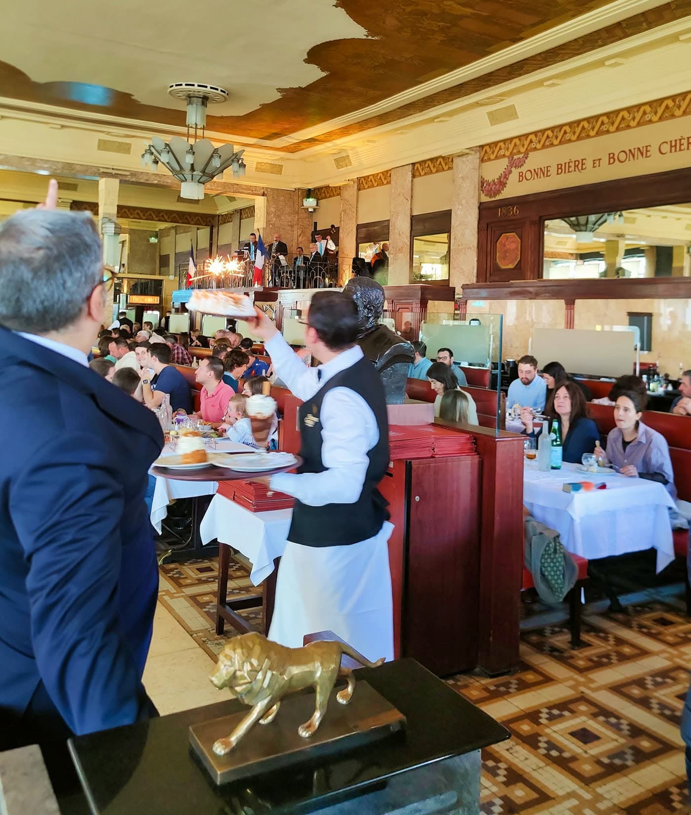 Serveur présente un gâteau d'anniversaire avec bougie allumée à une table de clients, statue de lion au premier plan — Brasserie Georges
