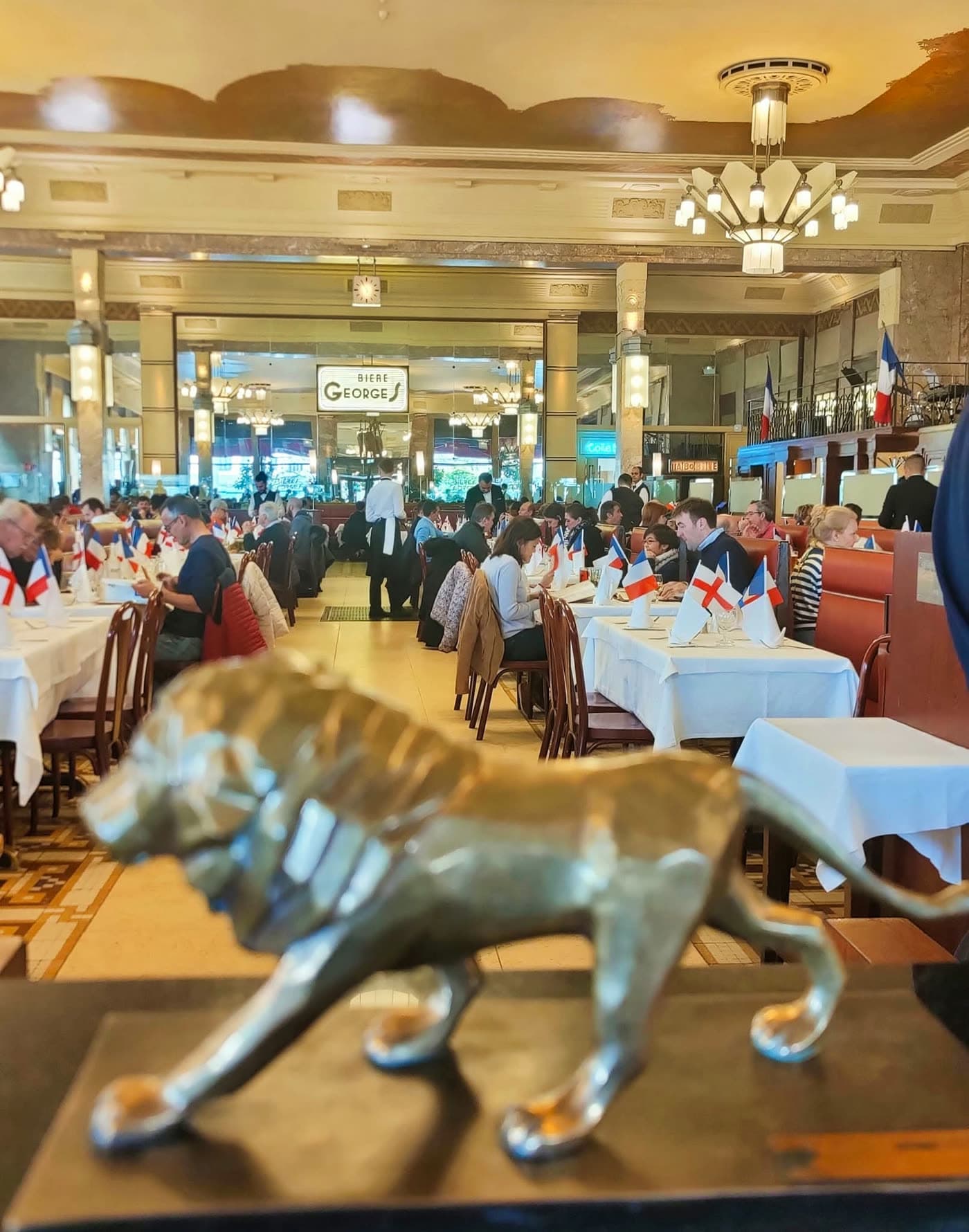 Statue de lion doré au premier plan devant salle animée avec petits drapeaux sur les tables — Brasserie Georges