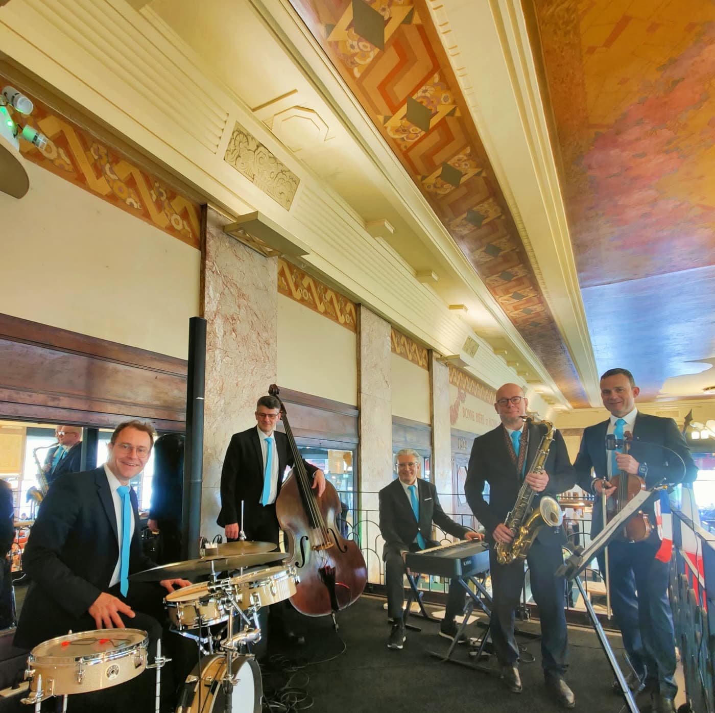 Groupe de jazz à cuivres (trompette, saxophone, contrebasse, batterie) joue sous la frise peinte du plafond — Brasserie Georges