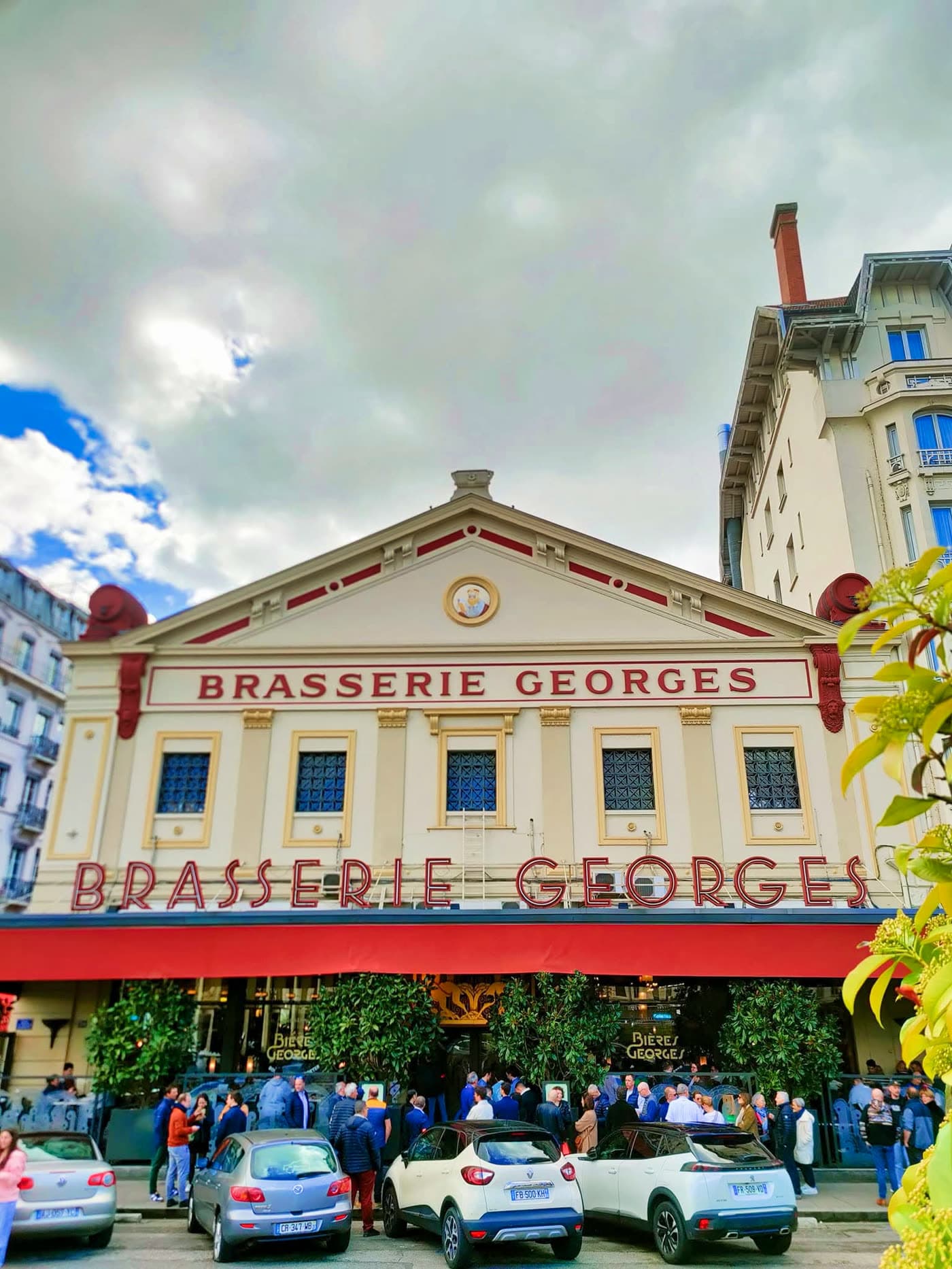 Façade Brasserie Georges en contre-plongée sous un ciel nuageux dramatique, voitures garées devant