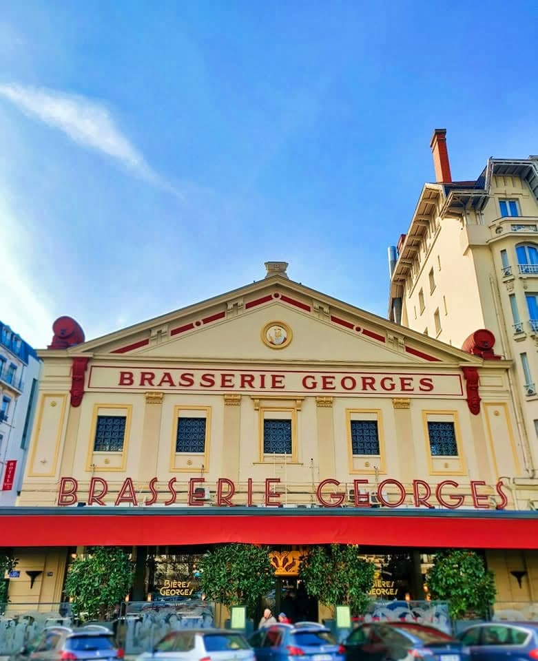 Façade Brasserie Georges sous ciel bleu, enseignes rouges bien visibles, voitures garées devant