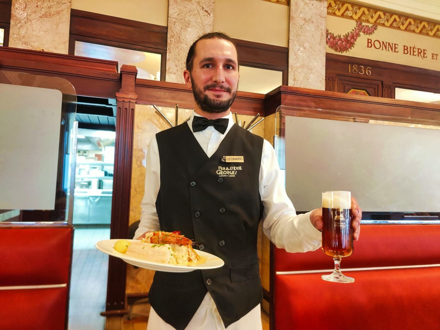 Serveur en gilet noir présente un plat garni et une chope de bière brune, salle en arrière-plan — Brasserie Georges