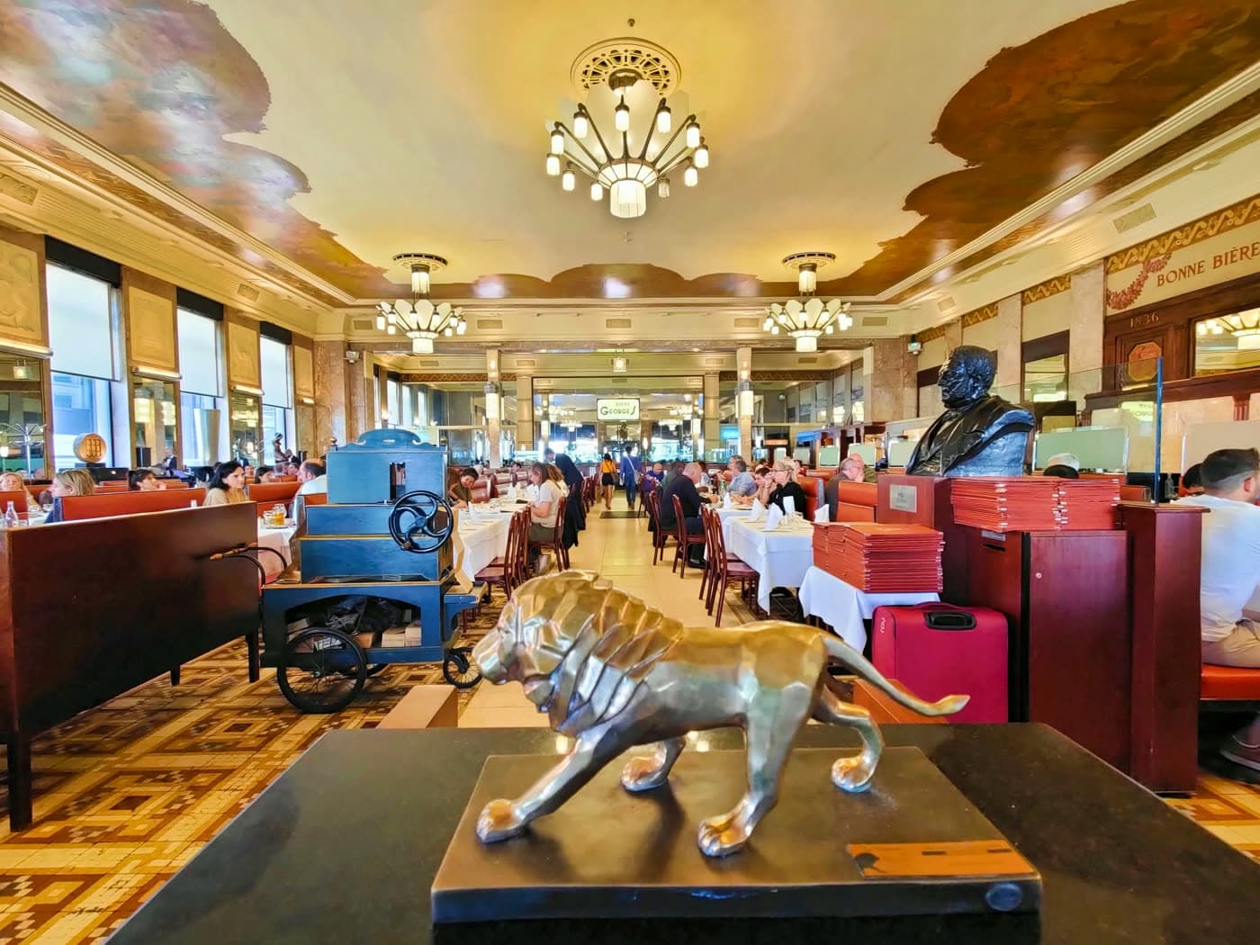 Vue de la salle Art déco avec chariot bleu, lion en bronze et plafond orné, clients attablés au fond — Brasserie Georges