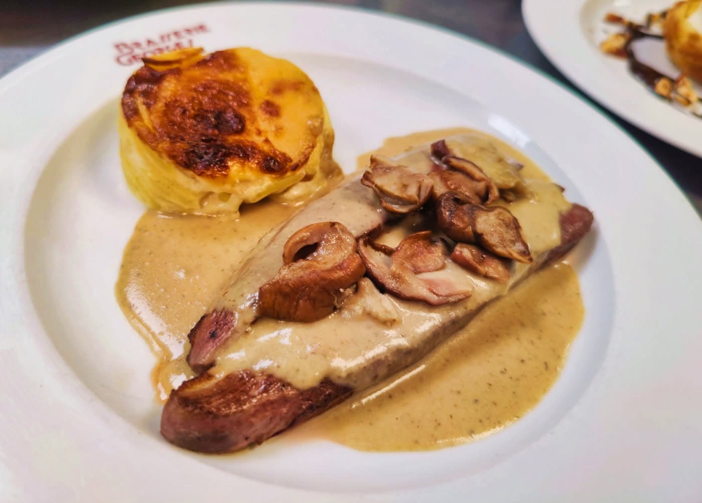 Pièce de viande en sauce aux champignons avec gratin dauphinois, assiette logotée — Brasserie Georges
