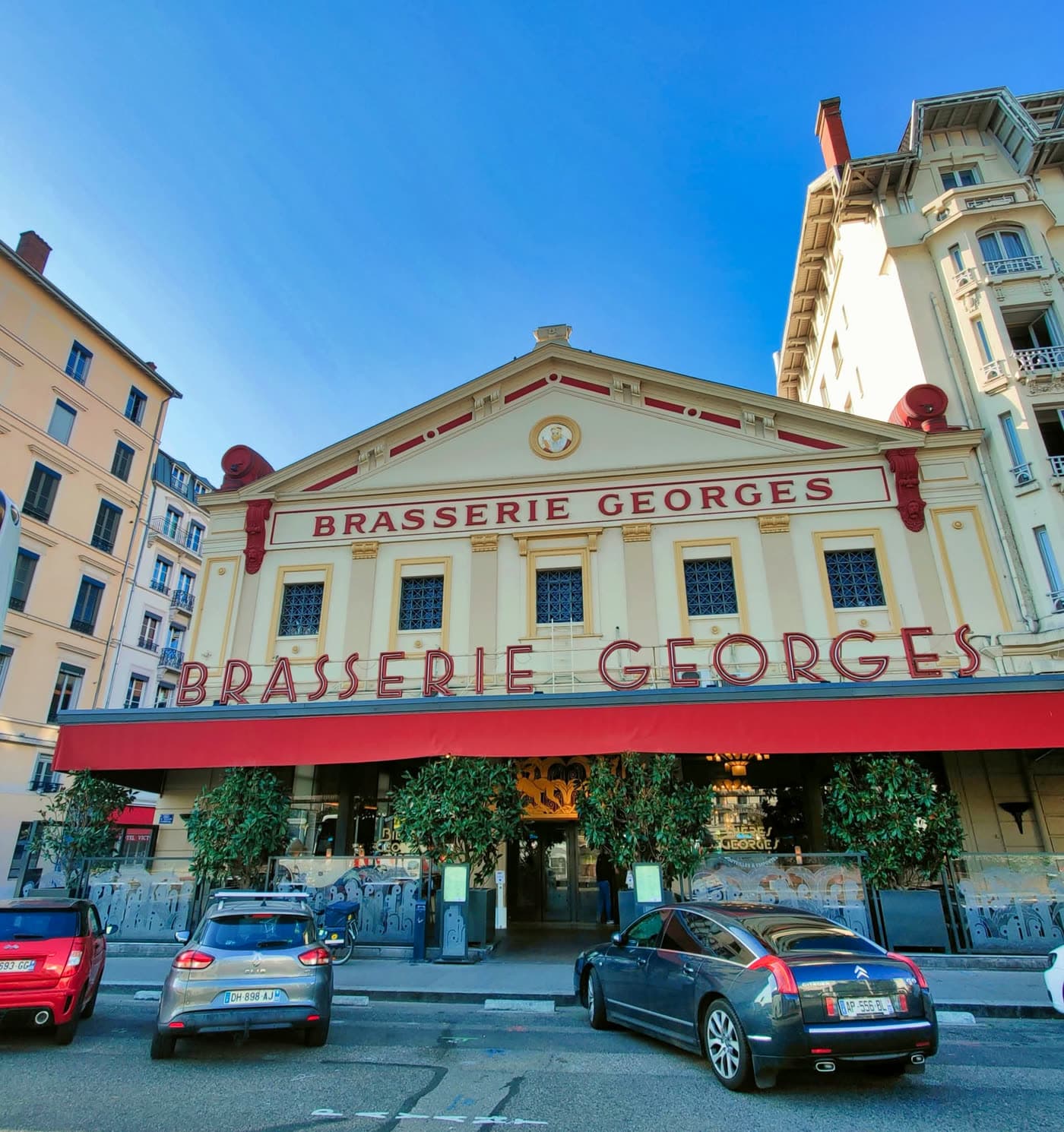 Façade Brasserie Georges de jour, auvent rouge, ciel bleu, voitures garées