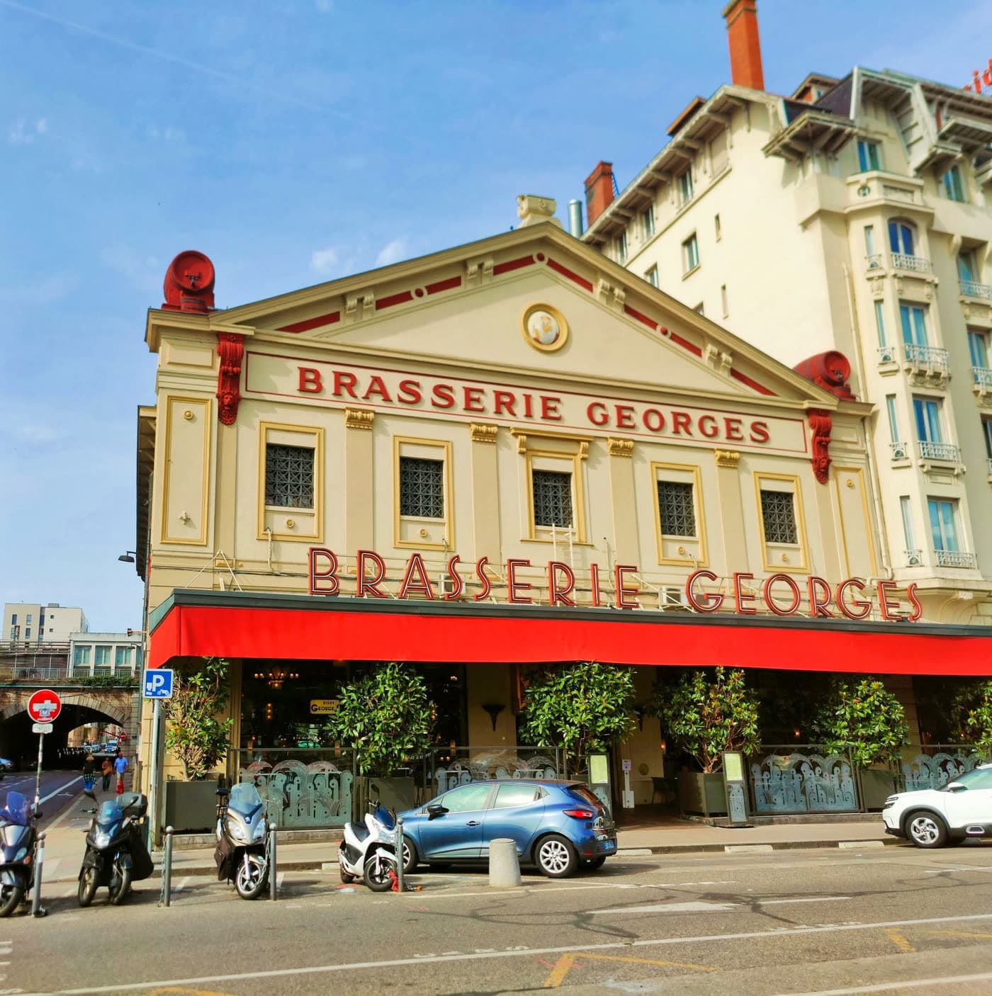 Façade grand angle avec auvent rouge et ciel bleu dégagé — Brasserie Georges