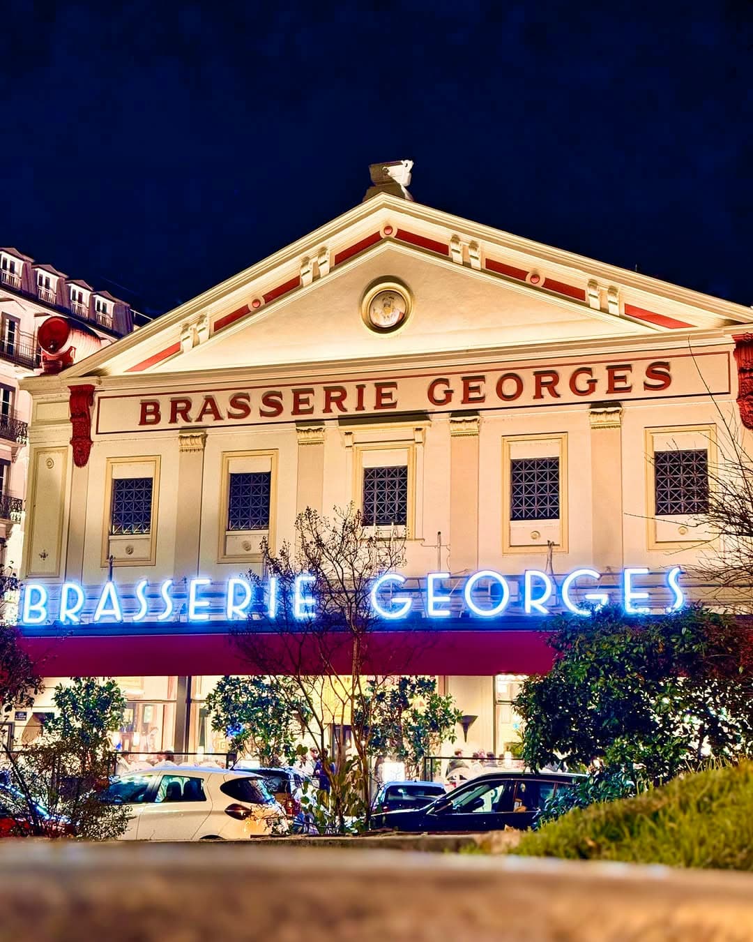 Façade illuminée la nuit avec enseigne néon bleue « Brasserie Georges » et fronton doré éclairé