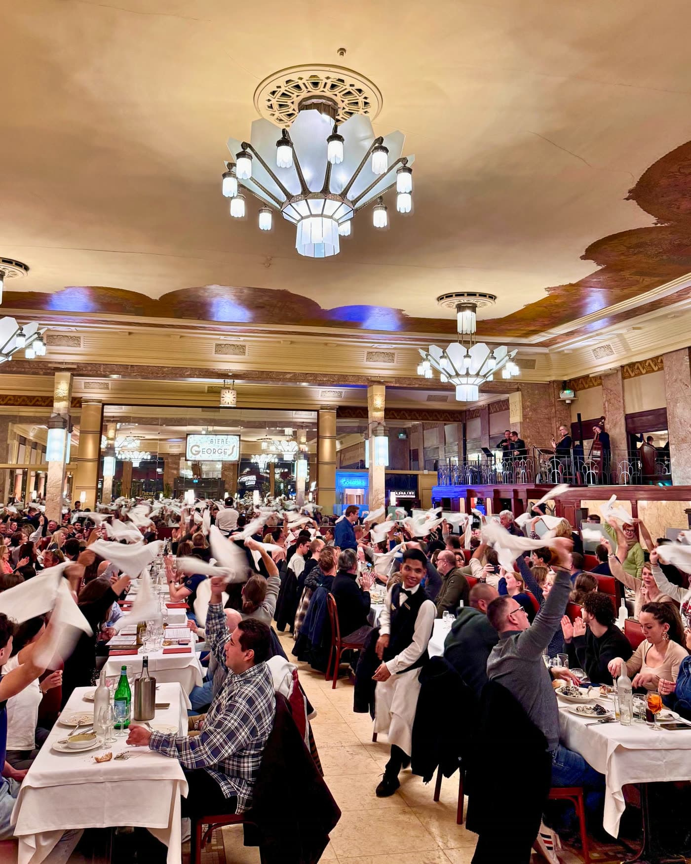 Convives agitant des serviettes blanches lors d'une animation festive, orchestre au fond sous les lustres — Brasserie Georges