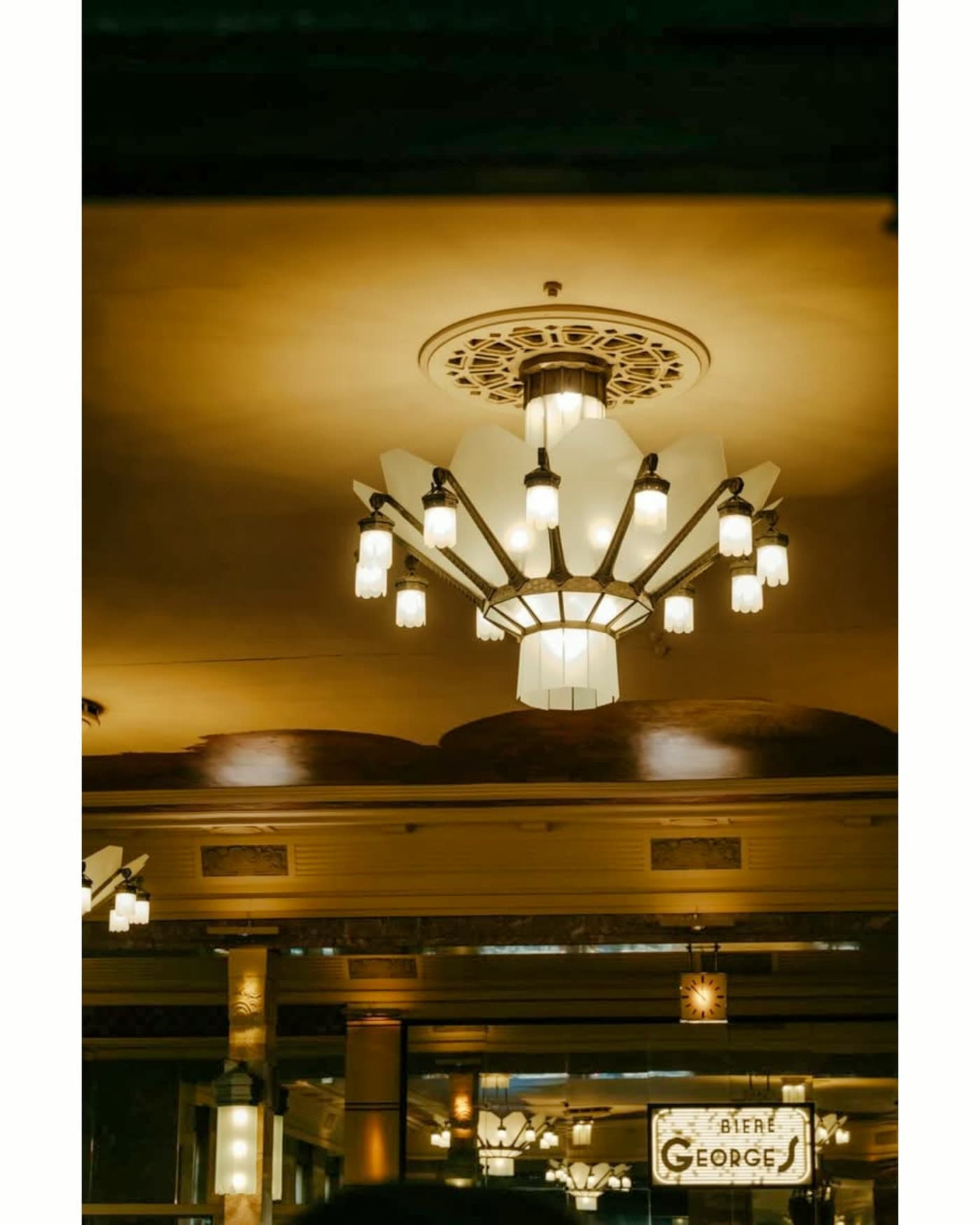 Plafond mouluré et grand lustre Art déco vu en contre-plongée, enseigne Bière Georges au fond — Brasserie Georges