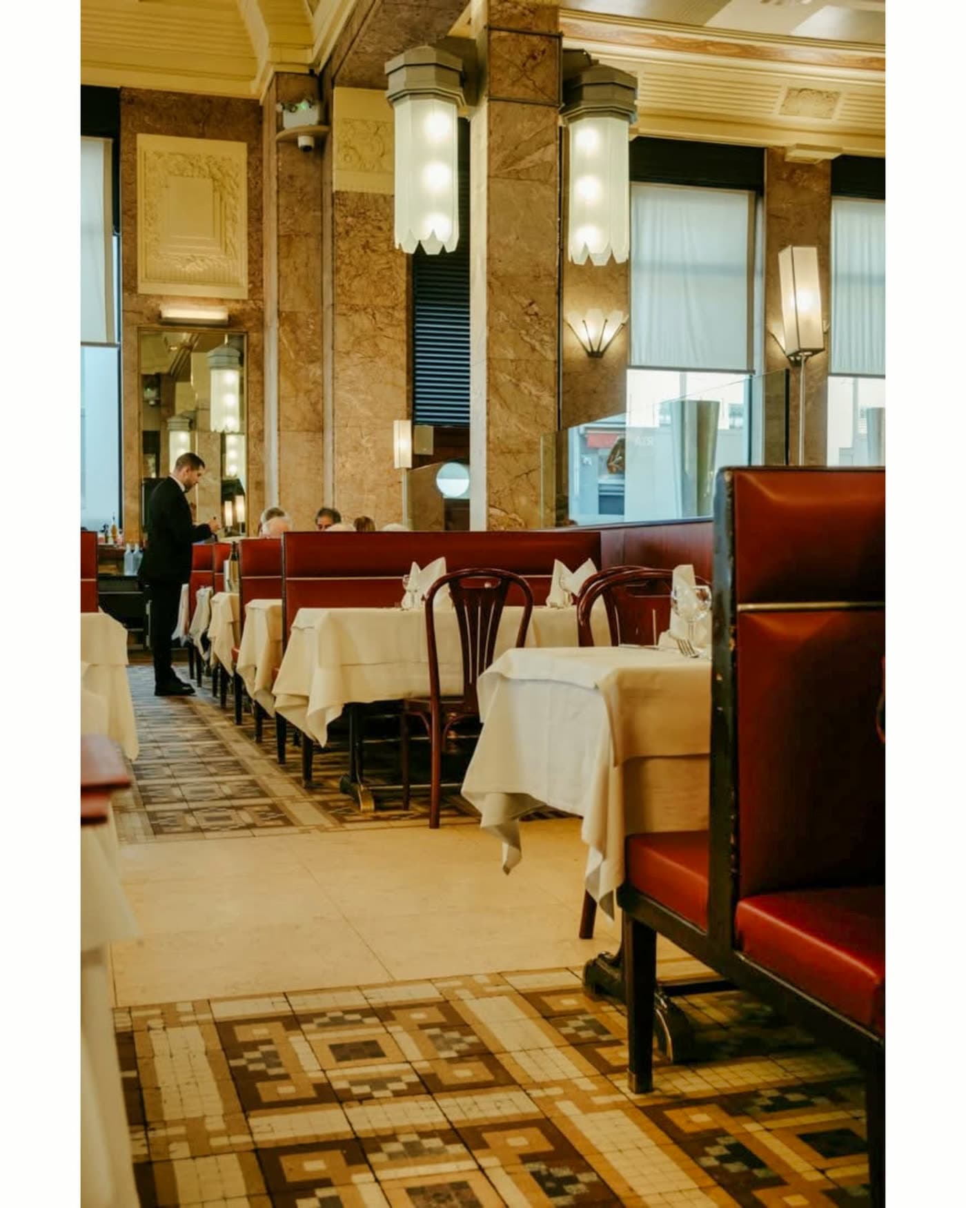 Colonnes en marbre, banquettes bordeaux et tables dressées, serveur au fond — Brasserie Georges