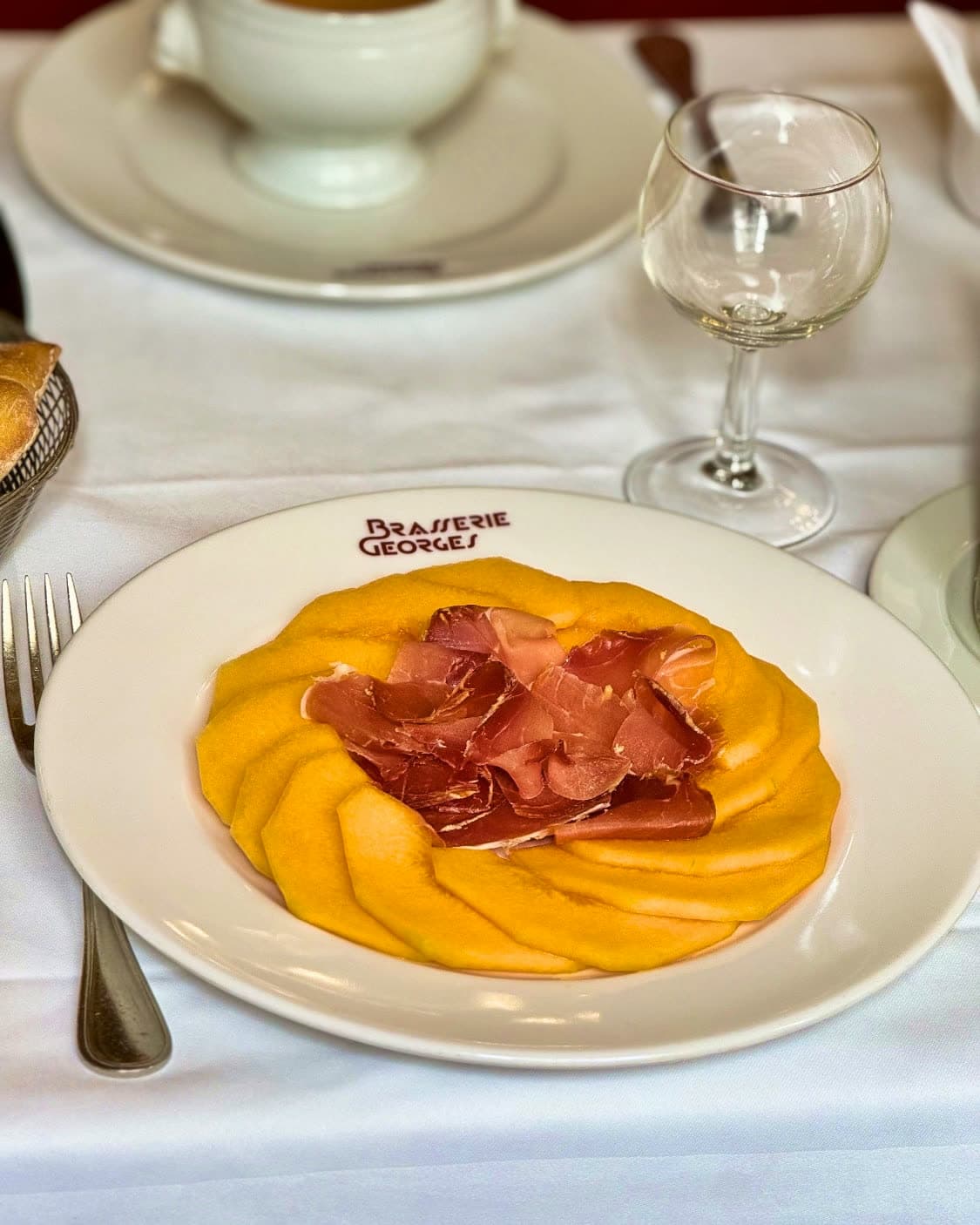 Assiette de melon et jambon cru sur vaisselle floquée Brasserie Georges, nappe blanche