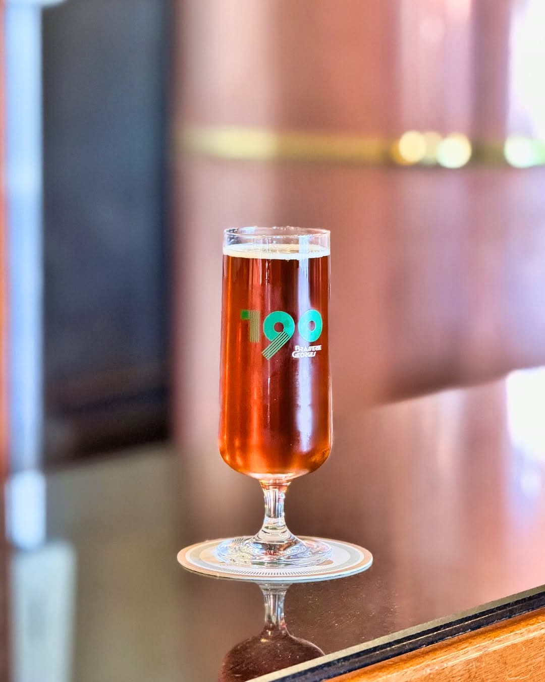 Verre de bière ambrée logotée '190' posé sur comptoir en laiton, reflets dorés — Brasserie Georges