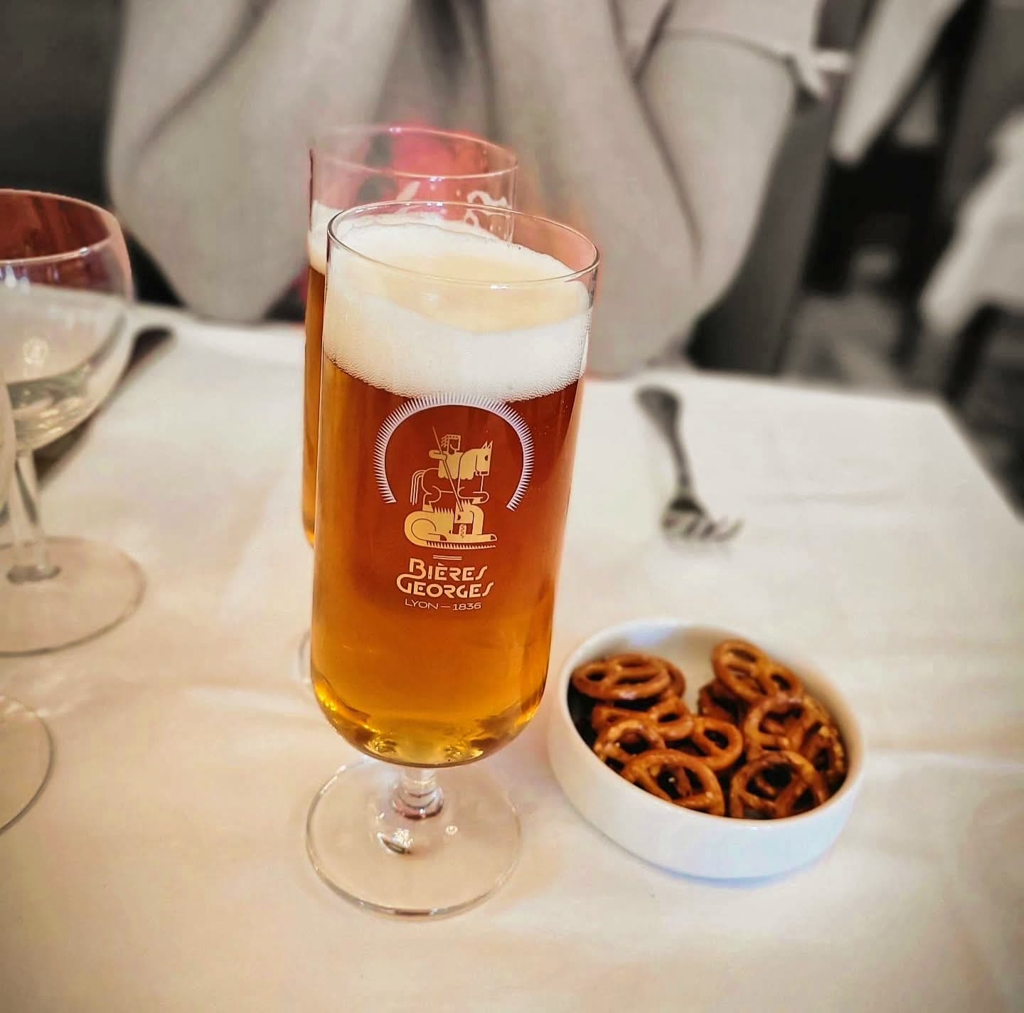 Un verre de bière Bières Georges servi avec son faux col, accompagné d'un bol de bretzels