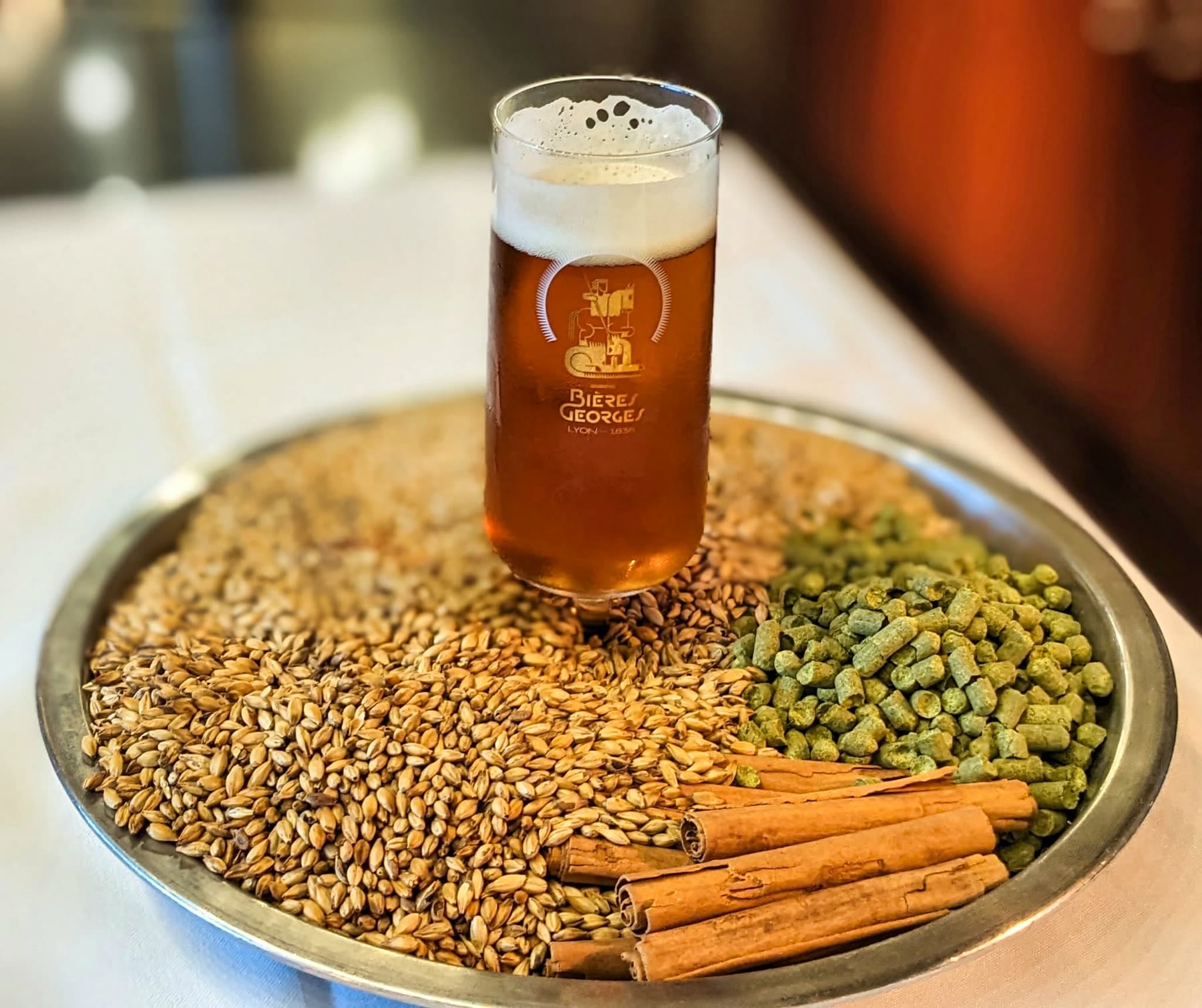 Un verre de bière Bières Georges posé sur un plateau de grains de malt, de cônes de houblon et de bâtons de cannelle